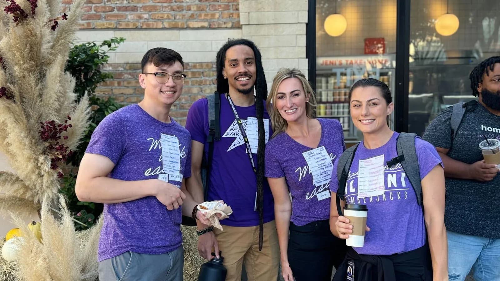 Group of four smiling people wearing purple shirts with name tags, standing outside near a brick building and holding drinks and snacks.