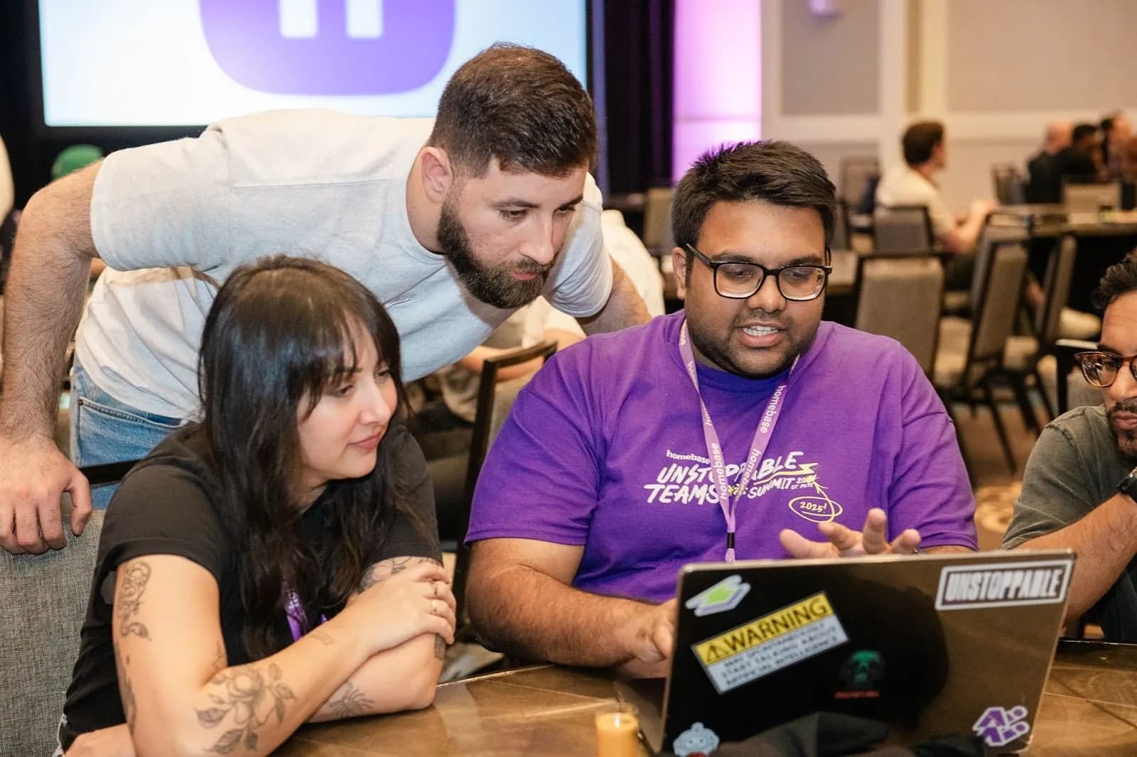Three people collaborating around a laptop, one in a purple shirt explaining something.
