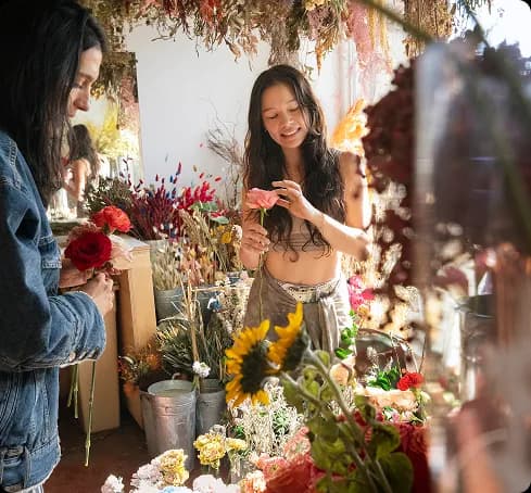 Two people arranging flowers in a florist shop filled with dried and fresh blooms, one holding a red rose and the other a pink flower.