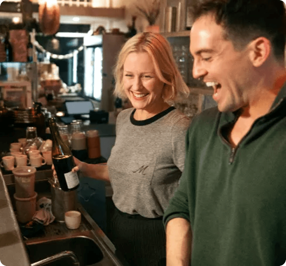 Two bar staff members smiling while working behind a counter at a restaurant or bar, representing small business owners using shift management software