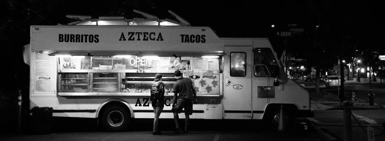Night scene of a food truck named Azteca serving burritos and tacos with two customers standing in front.