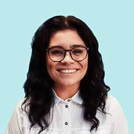 Smiling woman with long black hair, glasses, and a white collared shirt against a light blue background.