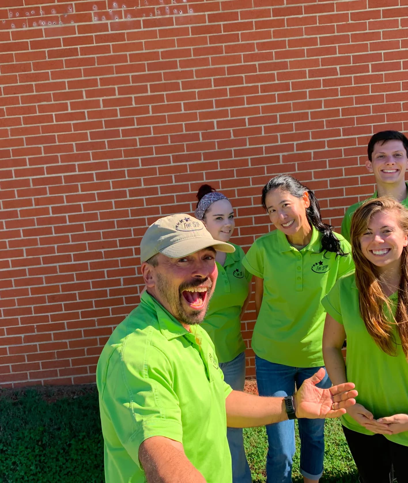 Five smiling people wearing matching bright green shirts posing cheerfully outdoors against a red brick wall.