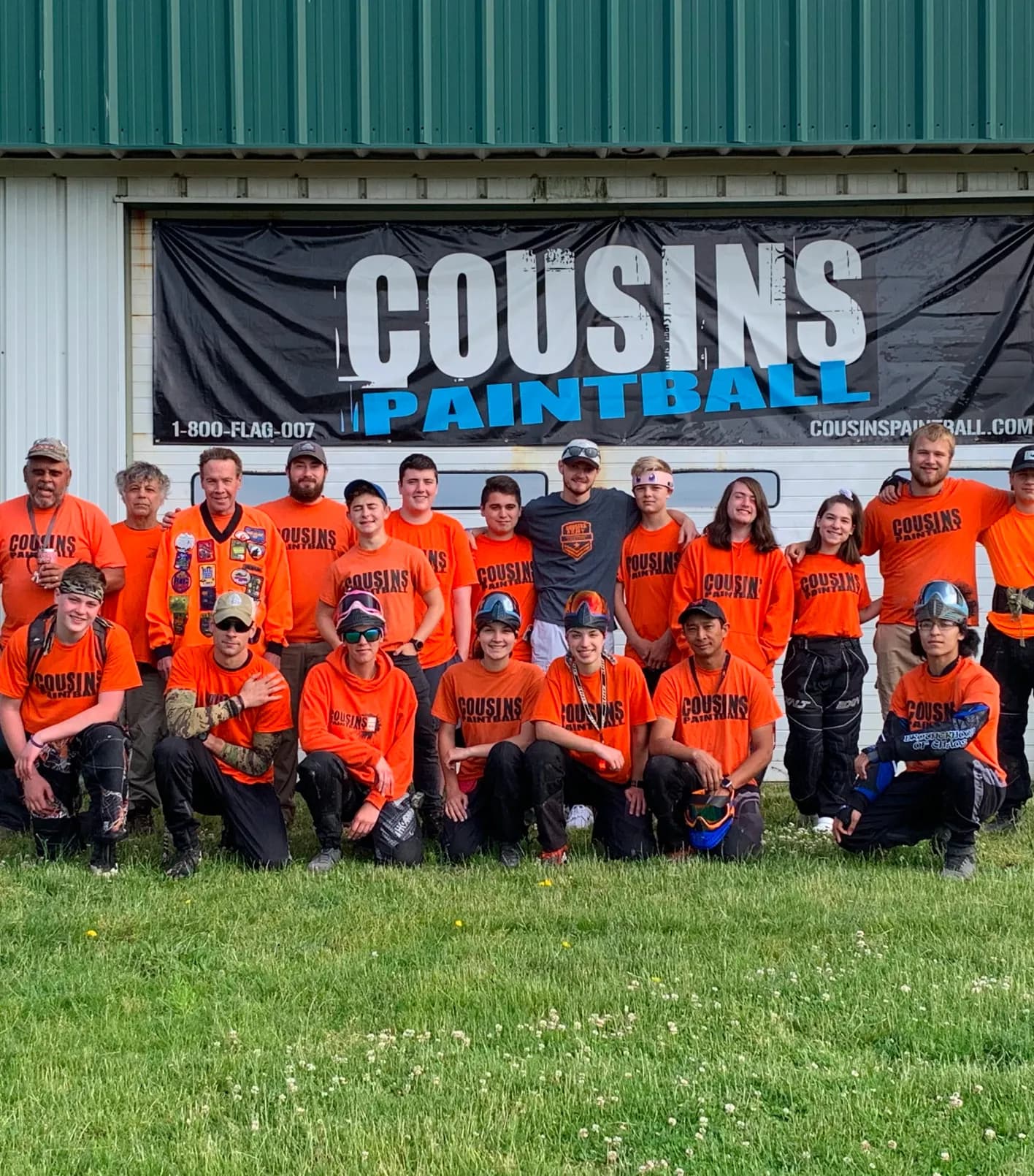 Group of people wearing orange Cousins Paintball shirts posing outdoors in front of a building with a Cousins Paintball banner.