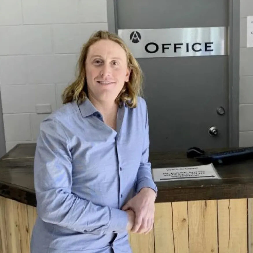 Smiling man with long blond hair wearing a blue shirt leaning against a wooden counter in front of an office door.