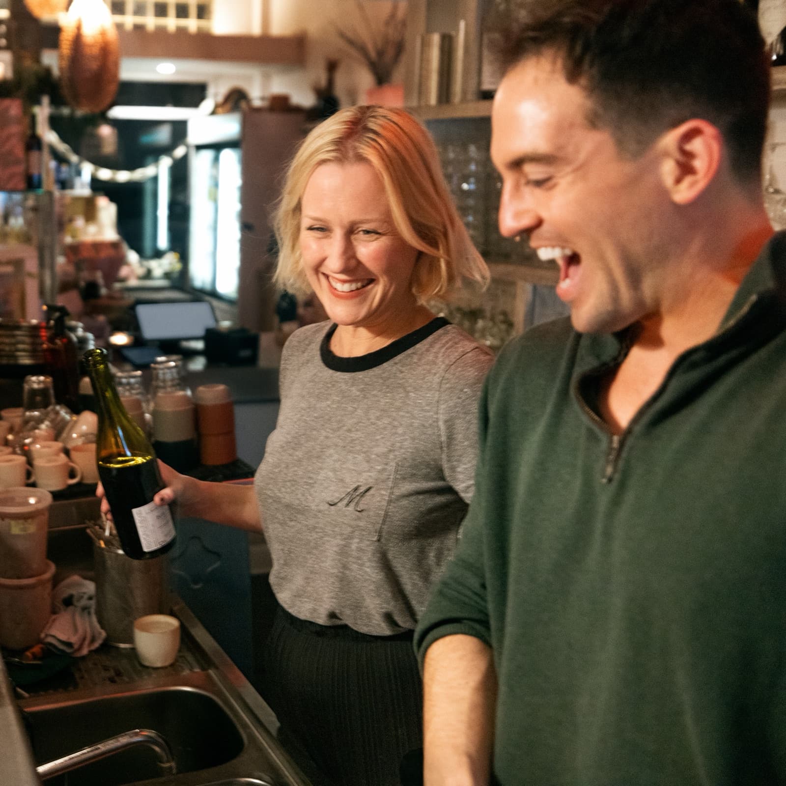 Two restaurant team members laughing behind the bar, one holding a bottle of wine.