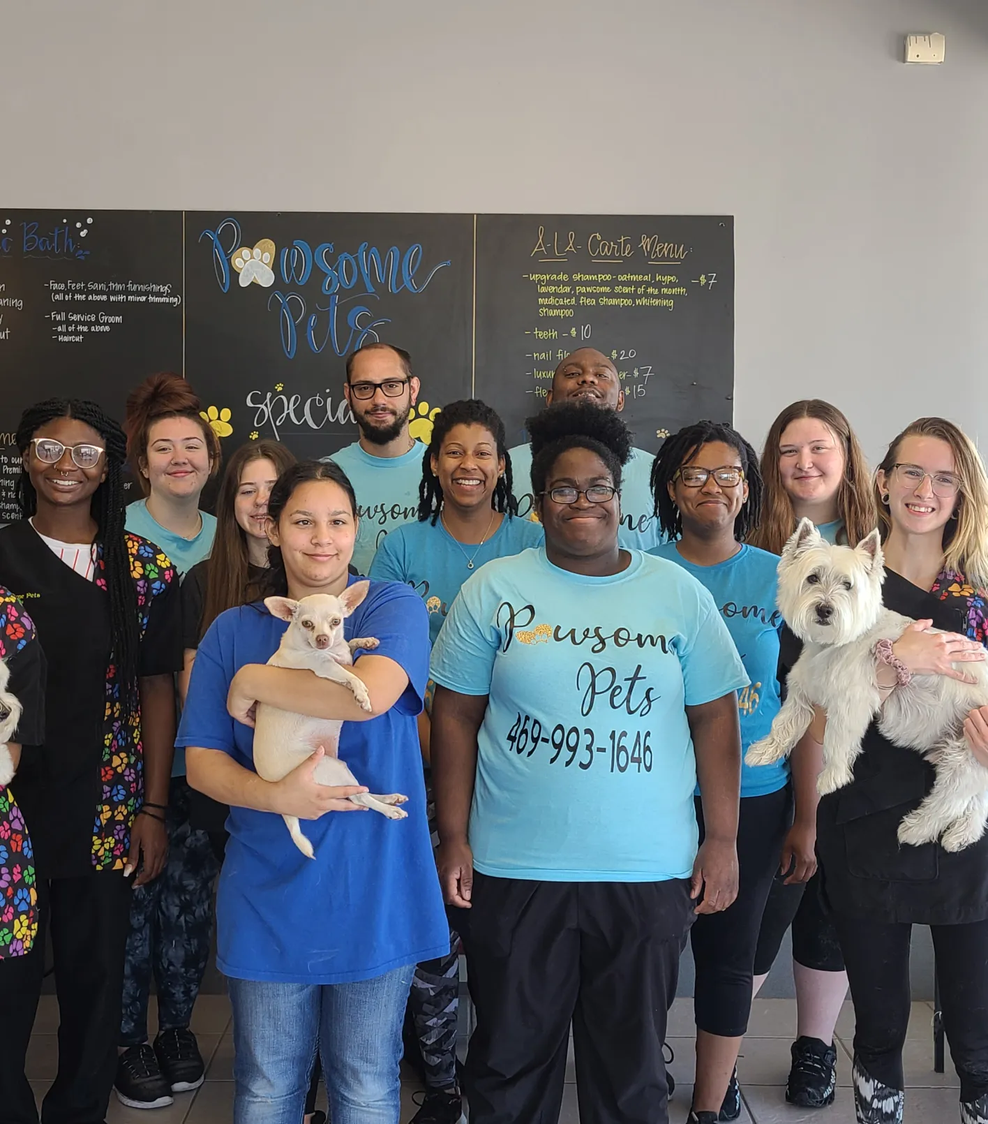 Group of smiling pet care staff posing indoors, some holding small dogs, with a blackboard menu in the background.