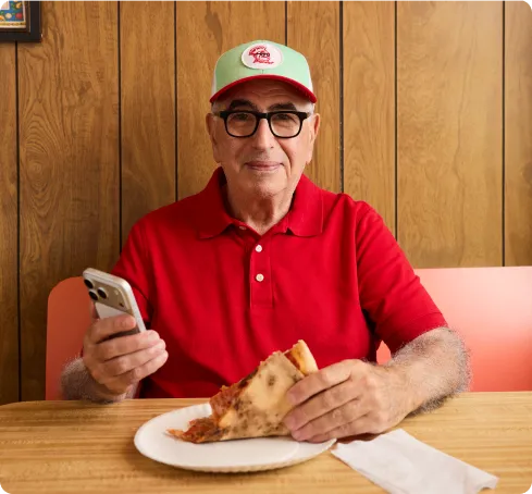 An older man in a red polo and cap sits at a wooden table. He smiles, holding a phone in one hand and a slice of pizza on a paper plate in the other..
