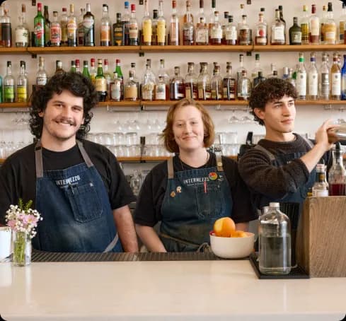 Three smiling bartenders stand behind a bar, wearing aprons. Shelves of colorful liquor bottles line the wall. A vase of flowers adds a cheerful touch.