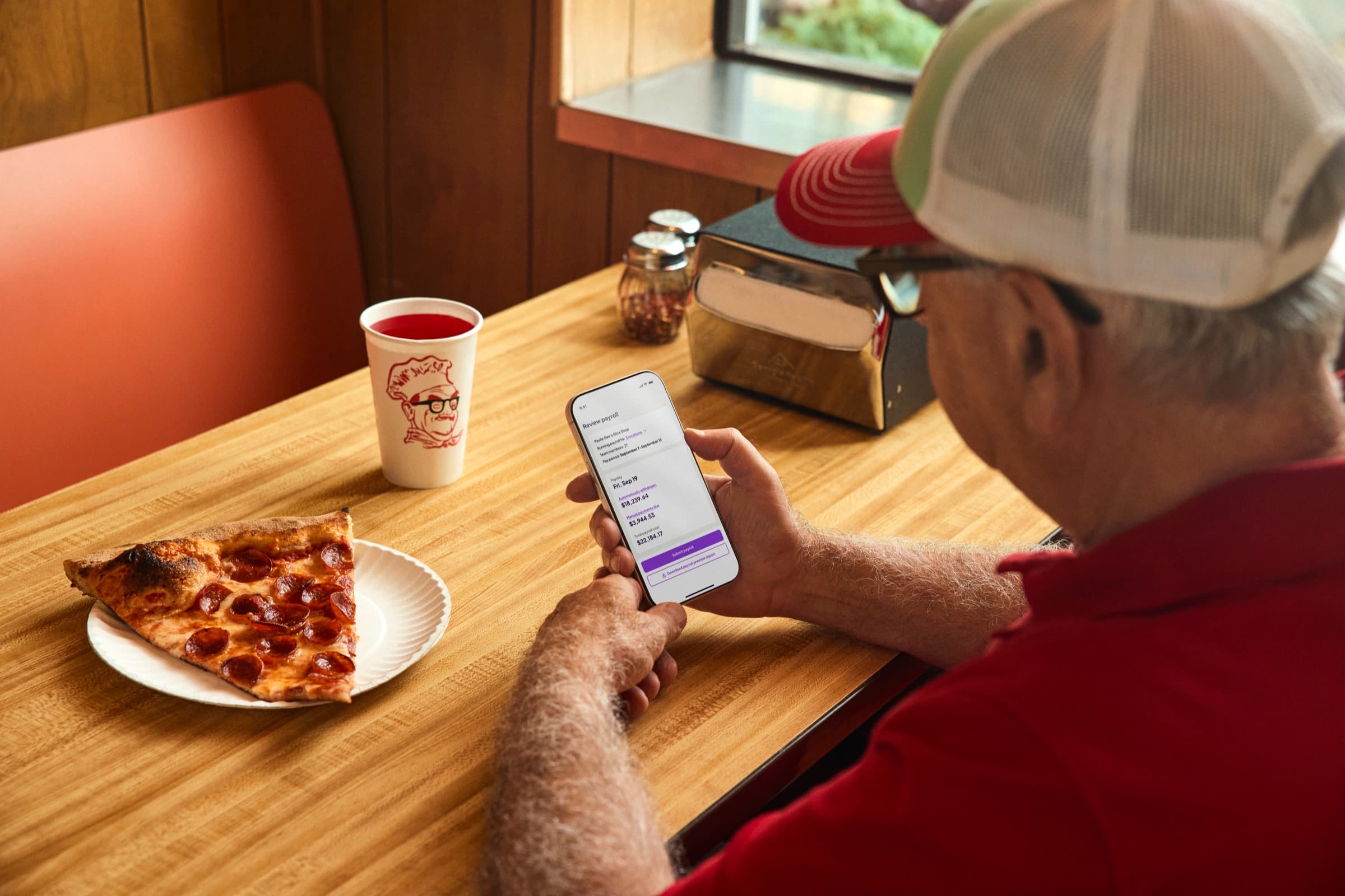 A pizzeria owner in a red shirt and trucker hat checking his phone beside a slice of pizza.
