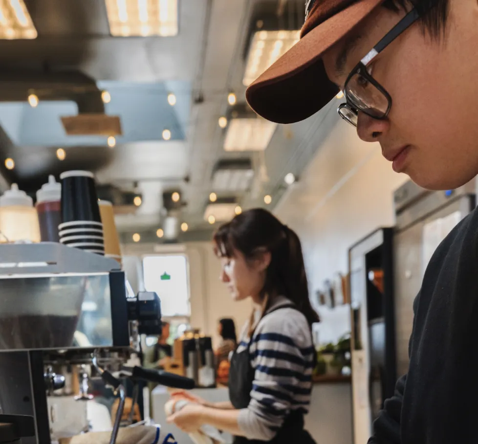 A young barista in a cap and glasses works at an espresso machine while a coworker prepares drinks behind them in a busy coffee shop.