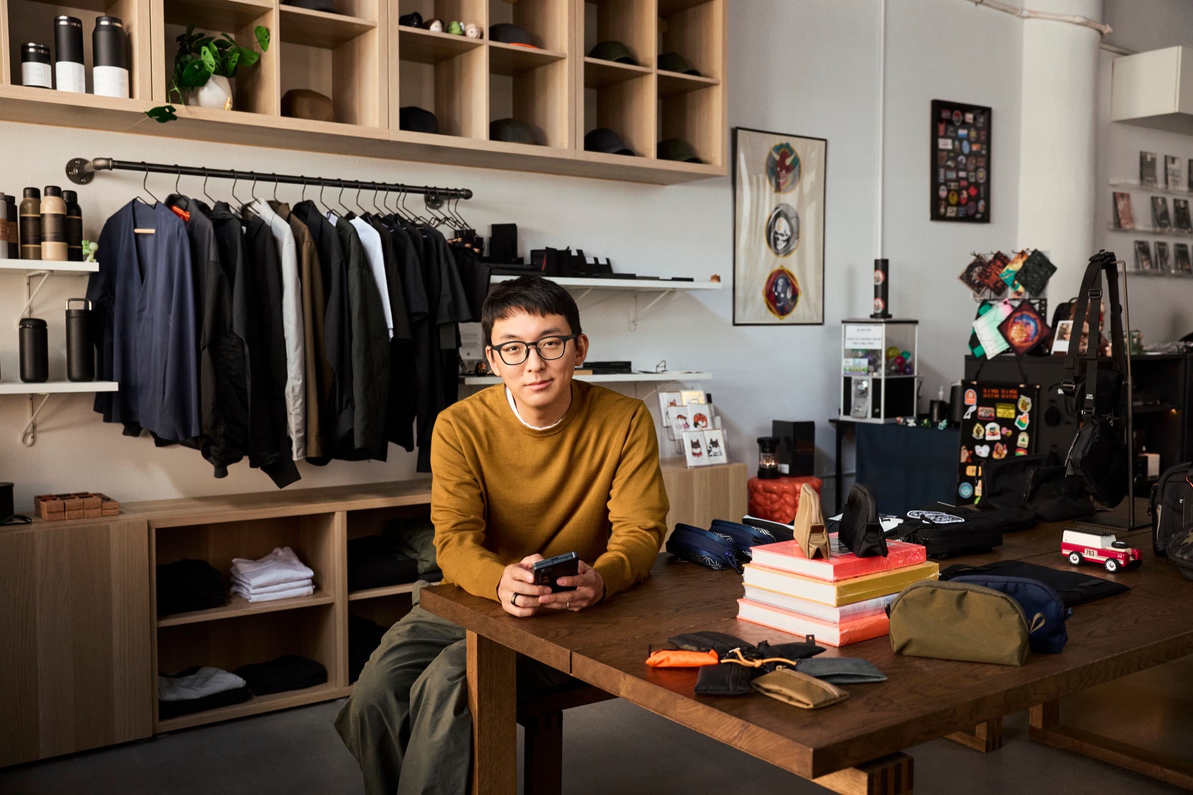 A shop owner sitting at his worktable holding a phone, surrounded by apparel and shelves.