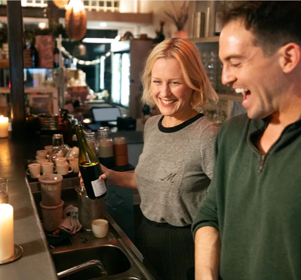 A smiling woman holds up a wine bottle beside a laughing man at a restaurant bar counter lit by candles.