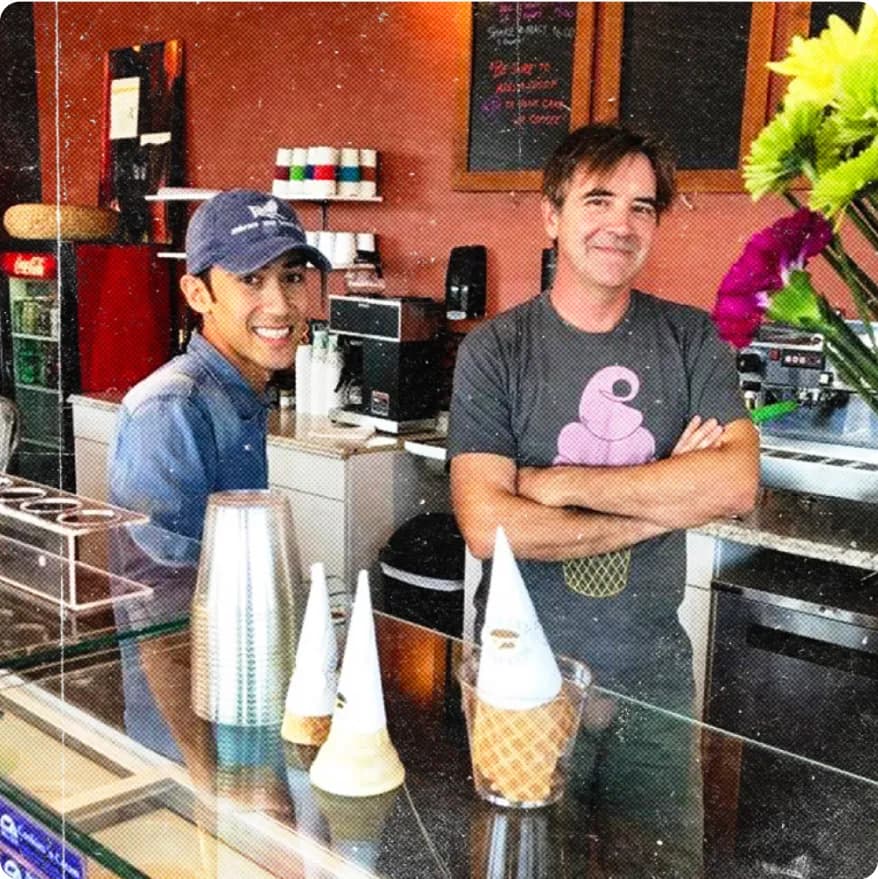 Two men smiling behind a cafe counter with soft serve ice cream cones displayed.