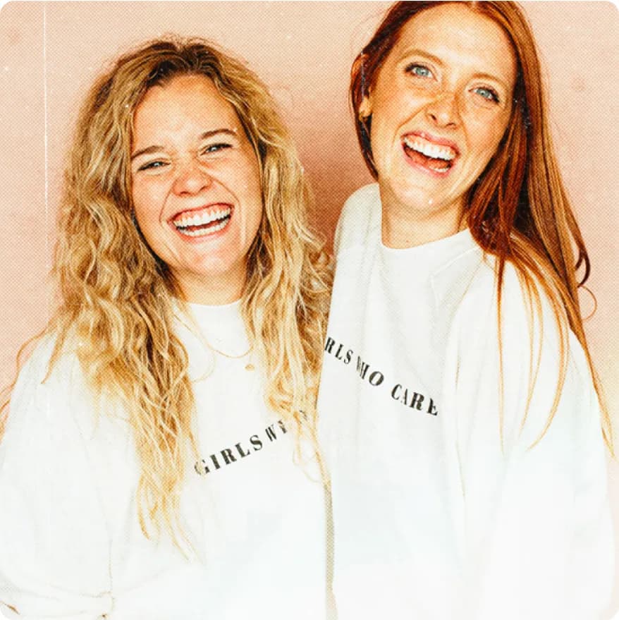 Two smiling women wearing white sweatshirts with 'GIRLS WHO CARE' printed on them, standing closely against a peach background.