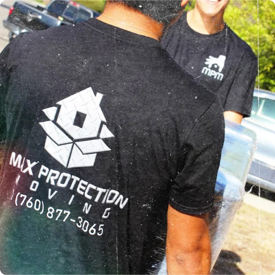 Two men wearing black Max Protection Moving t-shirts carrying a wrapped household item outdoors.