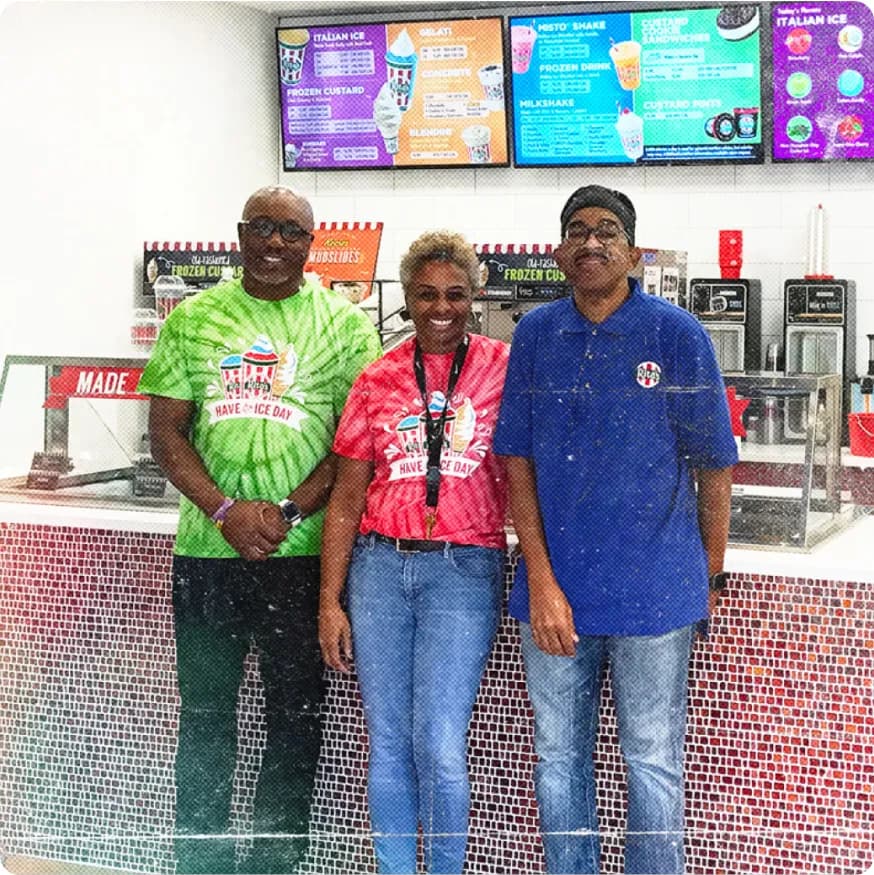 Three smiling Rita's employees standing behind the counter with colorful menu boards above them.