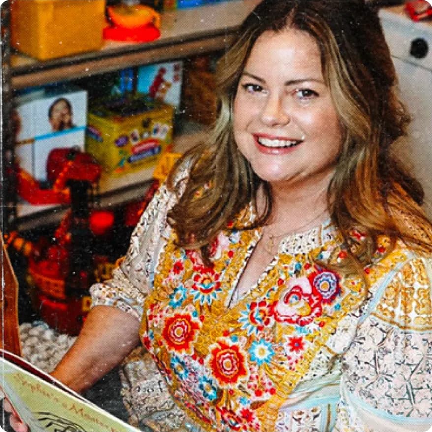 Smiling woman with long brown hair wearing a colorful embroidered top, seated in a room with shelves and various objects in the background.
