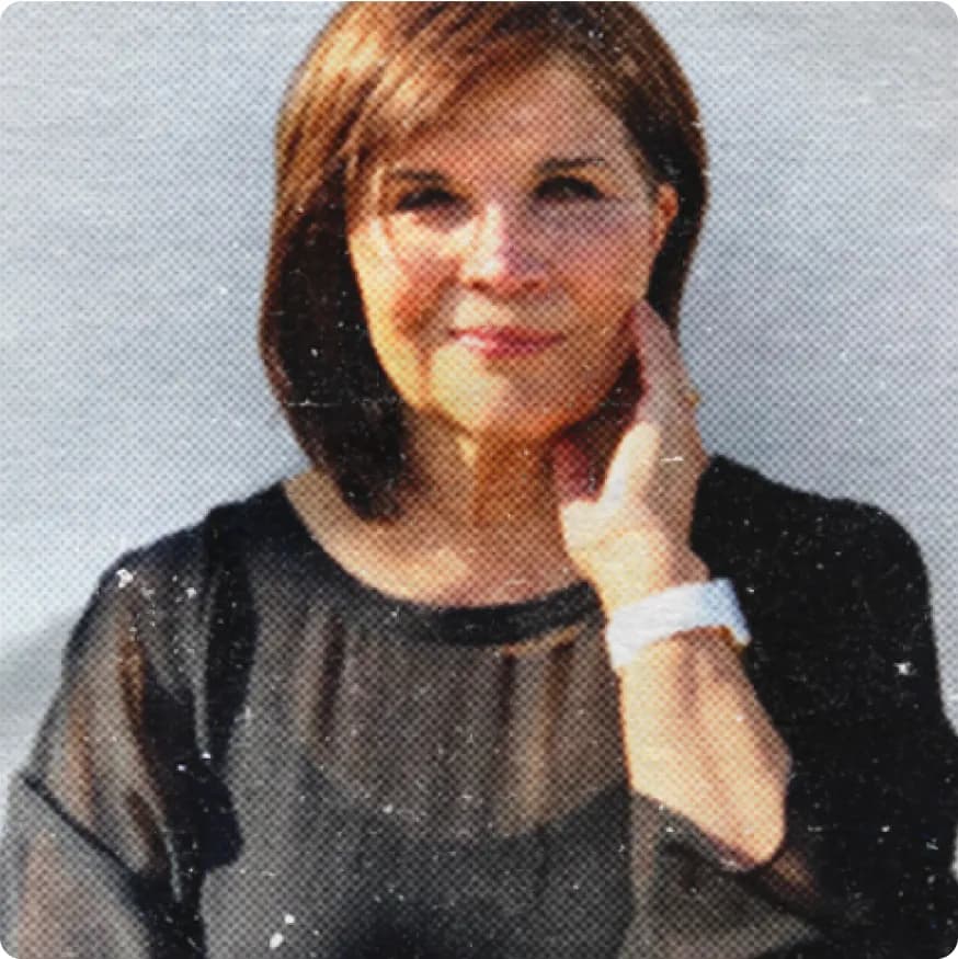 Middle-aged woman with short brown hair wearing a sheer black top and a white watch, touching her neck and smiling slightly.