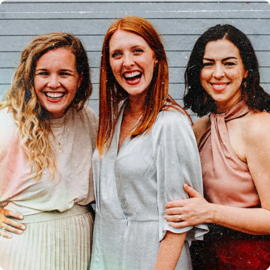 Three smiling women standing closely together in front of a gray wall.