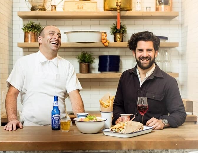 Two men smiling behind a wooden counter with food and drinks, one wearing a white shirt and apron, the other in a dark jacket, with shelves and plants in the background.
