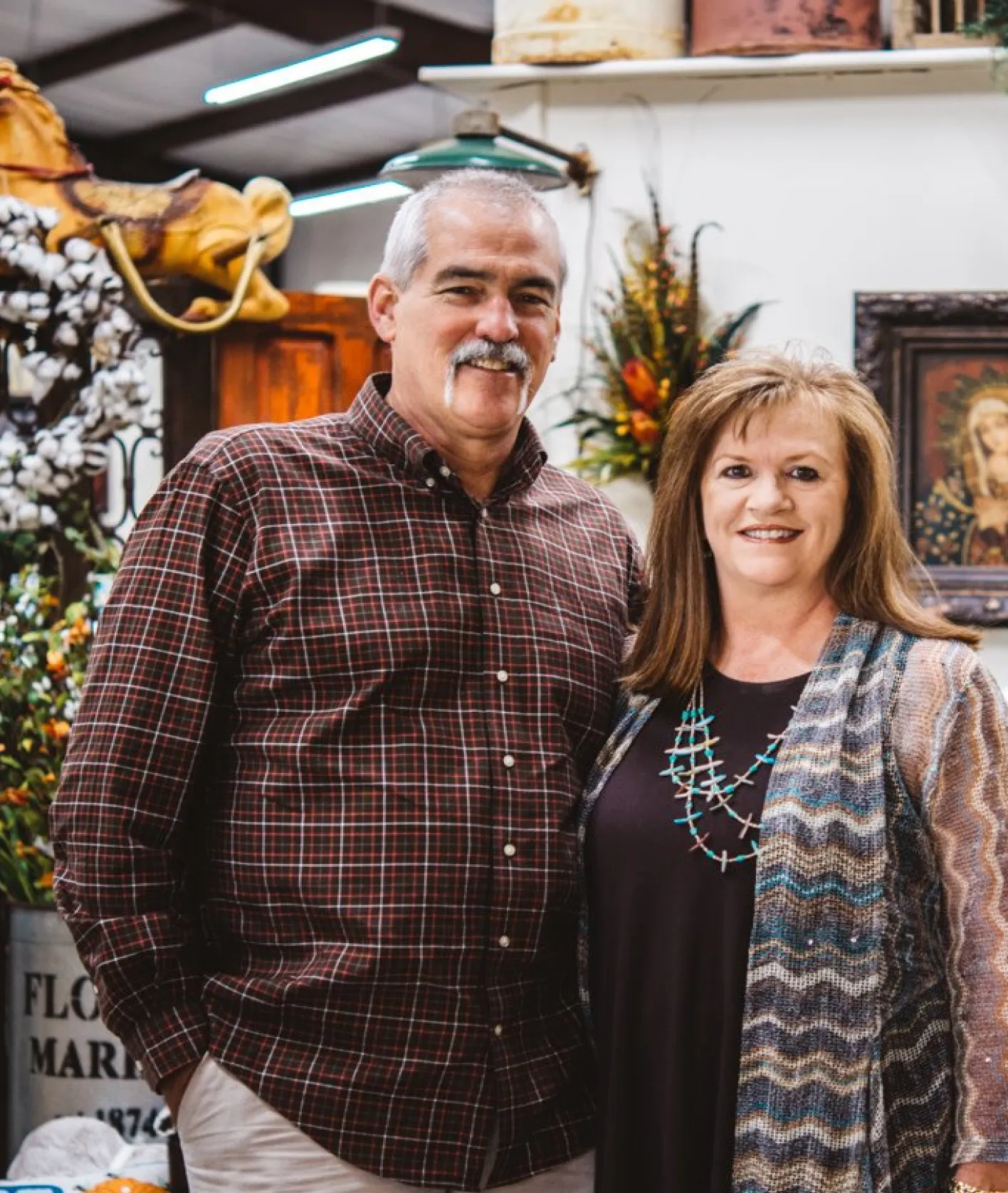 Smiling middle-aged man in a red plaid shirt and woman in a black dress with a patterned cardigan standing together indoors with floral decorations in the background.