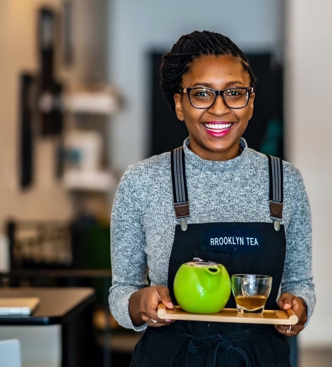 Smiling woman wearing glasses and a Brooklyn Tea apron holding a tray with a green teapot and a glass cup of tea.