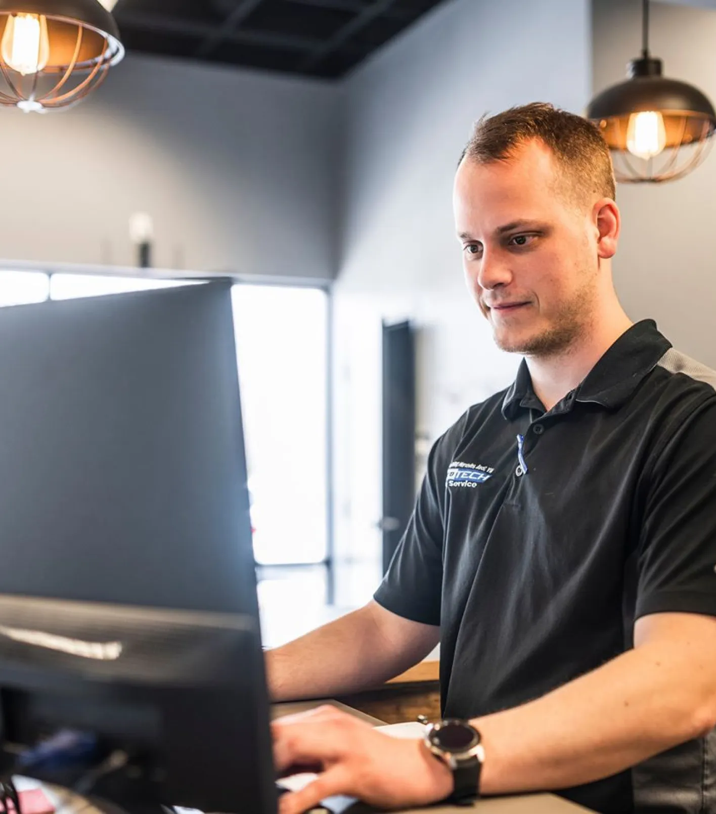 Man wearing a black polo shirt working on a computer in a modern office with hanging lantern lights.