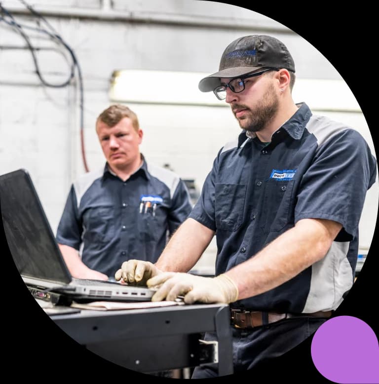Two mechanics in a workshop review a laptop on a rolling cart, one wearing safety glasses and gloves while typing.