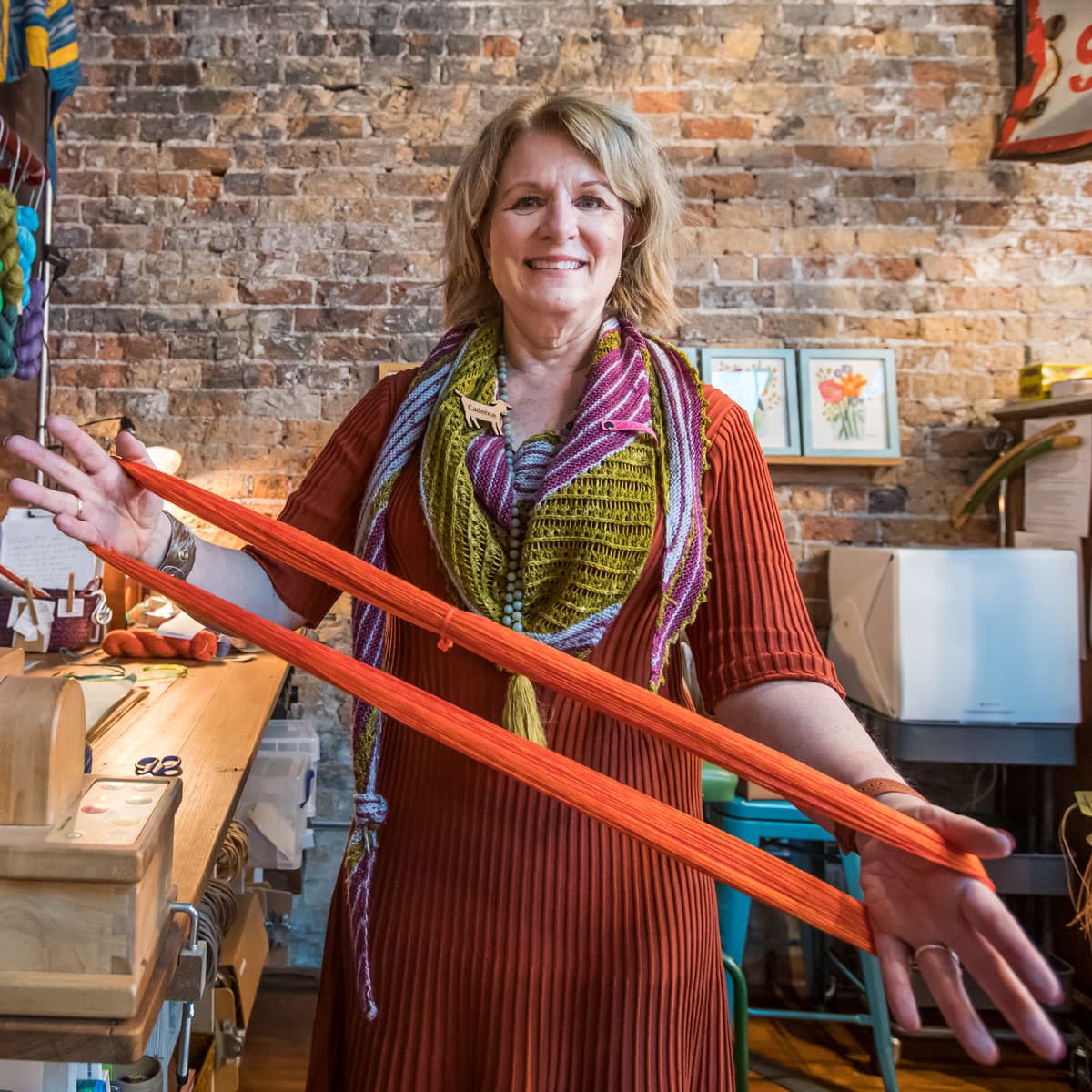 Smiling woman in a red dress and colorful scarf holding stretched orange yarn in a cozy craft studio with exposed brick wall.