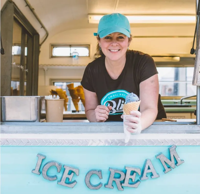 Smiling woman in a teal cap serving an ice cream cone from a pastel blue ice cream truck window.