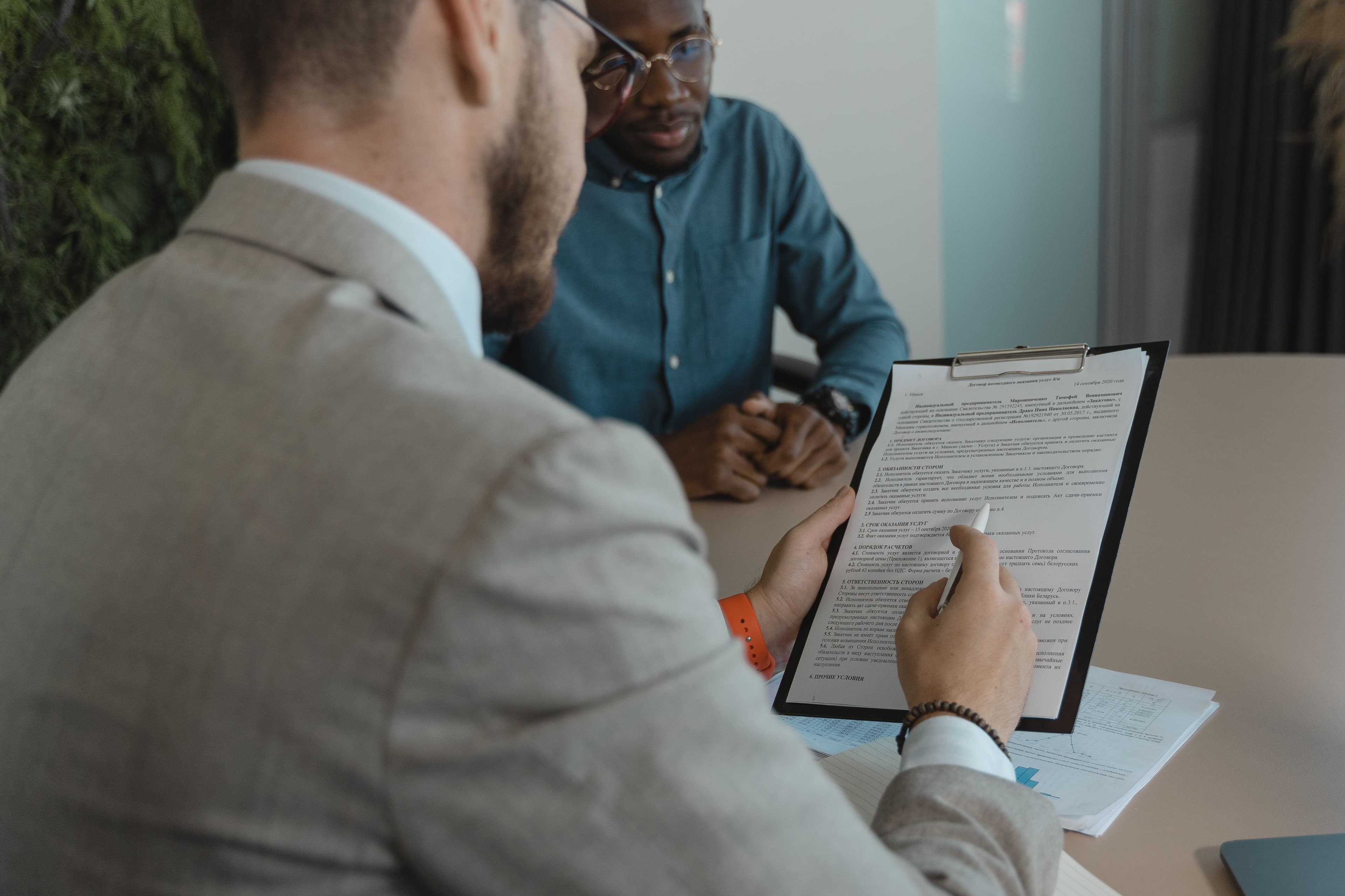 Two men sit across a table during a meeting as one, in a gray blazer, points a pen at a resume on a clipboard while the other listens.