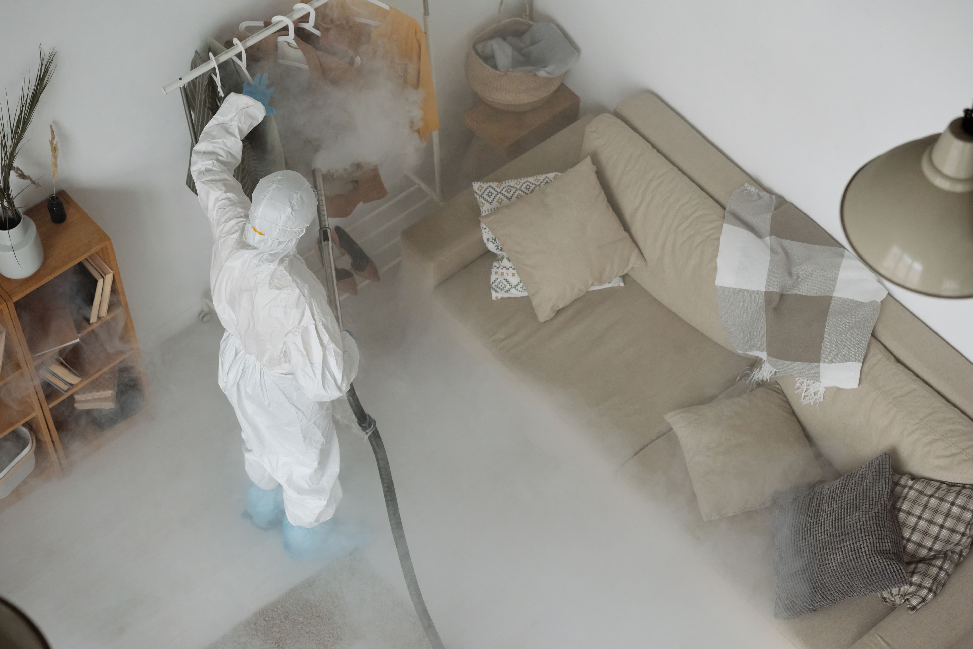Pest control technician in a white protective suit and shoe covers fogs a living room with a hose, as mist spreads across the floor near a beige sofa and clothing rack.