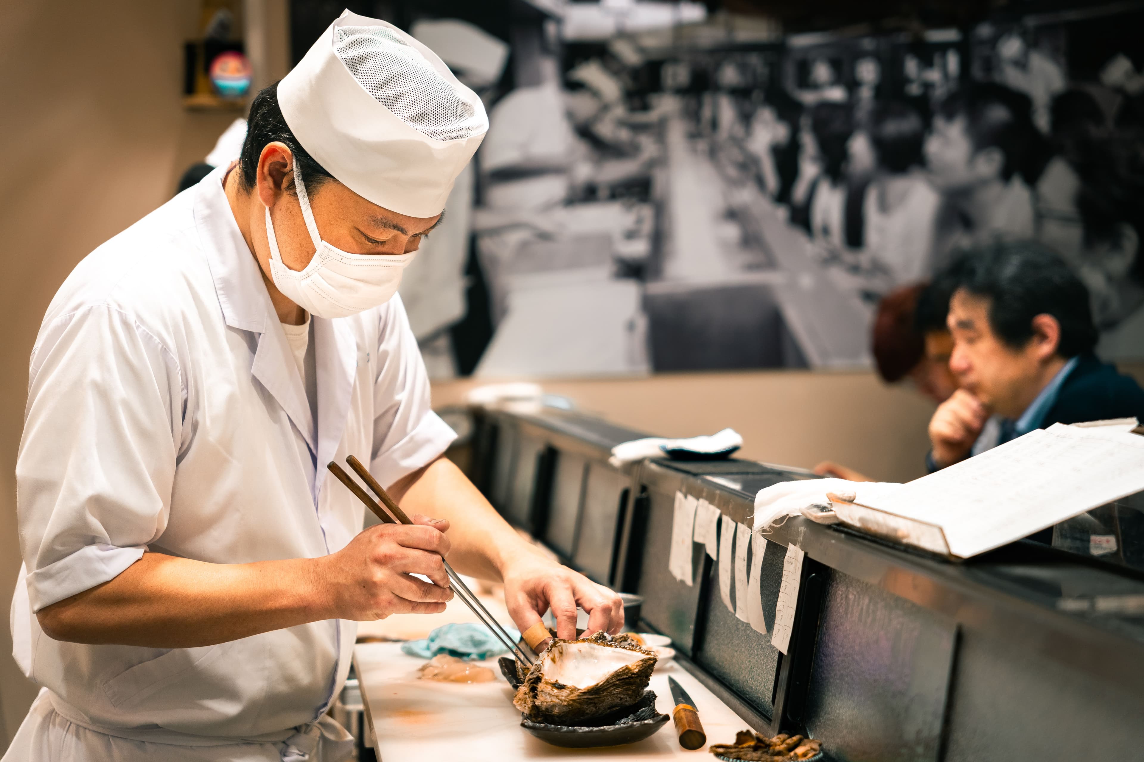 Sushi chef in a white uniform, cap, and mask uses chopsticks to prepare a dish at a restaurant counter while a customer sits in the background.