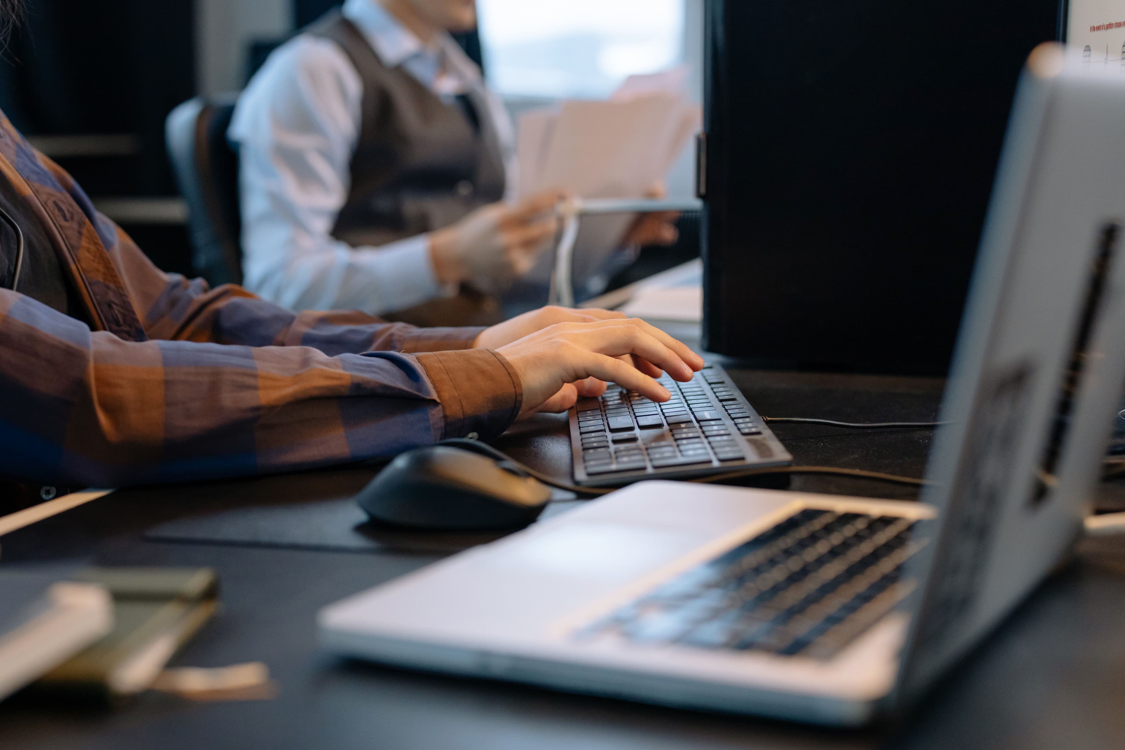 Close-up of a person in a plaid shirt typing on a desktop keyboard in an office, with a laptop in the foreground and a coworker holding papers in the background.